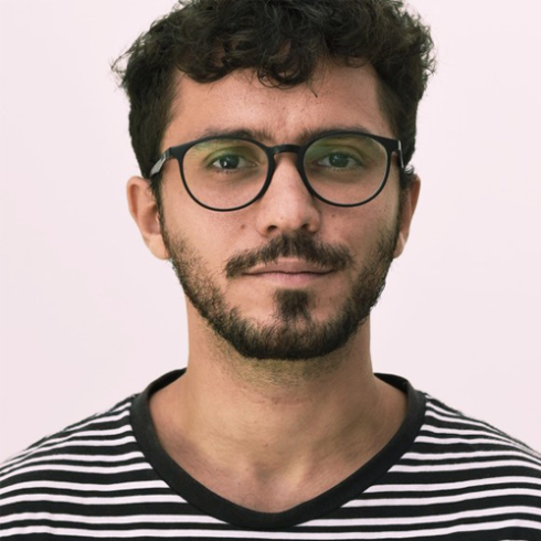 Lucas Almeida é moreno, tem cabelo escuro e barba, usa óculos e veste uma blusa com listras.